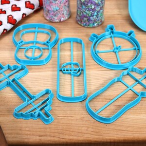May include: A set of six blue plastic cookie cutters shaped like a dumbbell, a protein shaker, a stopwatch, a weight, a protein bar, and a water bottle.