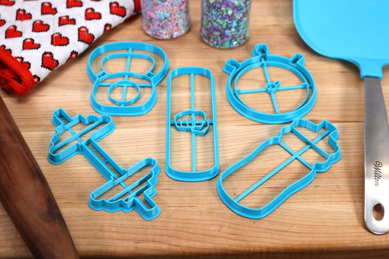 May include: A set of six blue plastic cookie cutters shaped like a dumbbell, a protein shaker, a stopwatch, a weight, a protein bar, and a water bottle.