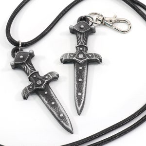 May include: Two silver-colored dagger pendants on black cords. One dagger is attached to a silver clasp.
