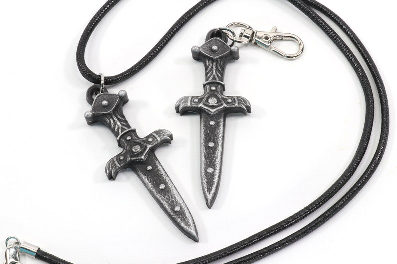 May include: Two silver-colored dagger pendants on black cords. One dagger is attached to a silver clasp.