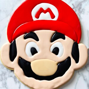 K&ouml;nnte beinhalten: Ein dekorierter Zuckerkeks in Form des Gesichts einer Videospielfigur. Der Keks ist mit rotem, wei&szlig;em und schwarzem Zuckerguss verziert. Die Figur tr&auml;gt einen roten Hut mit einem wei&szlig;en "M" darauf, schwarze Augenbrauen, wei&szlig;e Augen, einen schwarzen Schnurrbart und eine braune Nase.