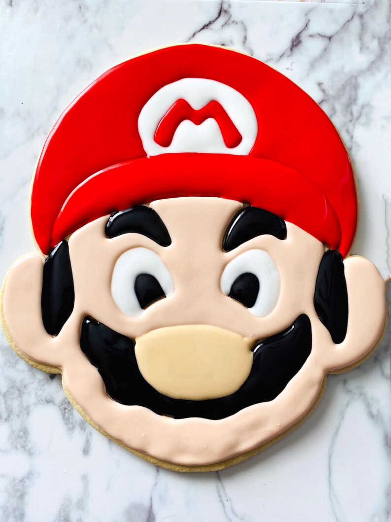 K&ouml;nnte beinhalten: Ein dekorierter Zuckerkeks in Form des Gesichts einer Videospielfigur. Der Keks ist mit rotem, wei&szlig;em und schwarzem Zuckerguss verziert. Die Figur tr&auml;gt einen roten Hut mit einem wei&szlig;en "M" darauf, schwarze Augenbrauen, wei&szlig;e Augen, einen schwarzen Schnurrbart und eine braune Nase.