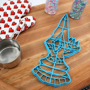 May include: Blue plastic cookie cutter shaped like a hand holding a large, round object. The cutter is on a wooden surface with a rolling pin, measuring cup, and sprinkles.