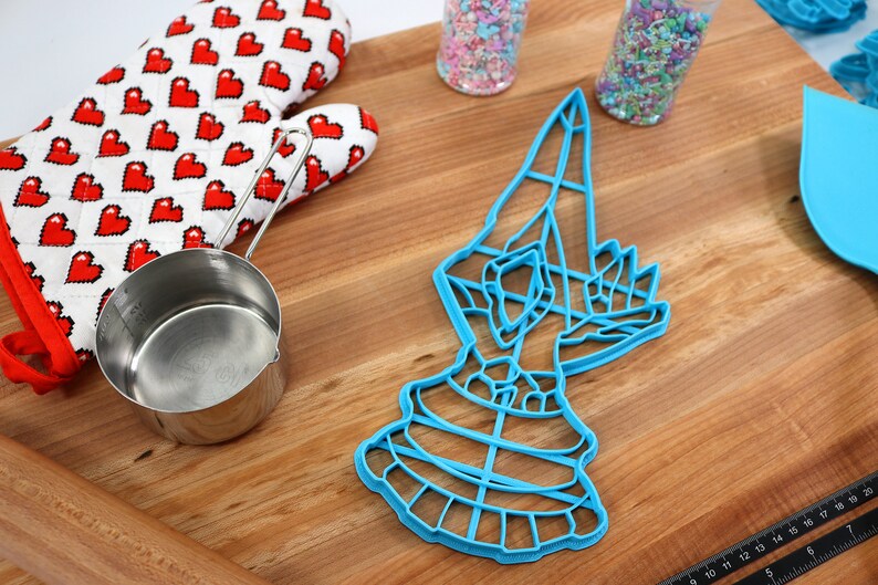 May include: Blue plastic cookie cutter shaped like a hand holding a large, round object. The cutter is on a wooden surface with a rolling pin, measuring cup, and sprinkles.