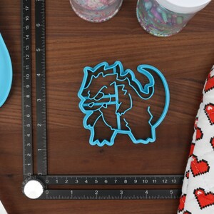 May include: A blue plastic cookie cutter shaped like a cartoon dragon. The dragon is curled up with its head resting on its front paws.