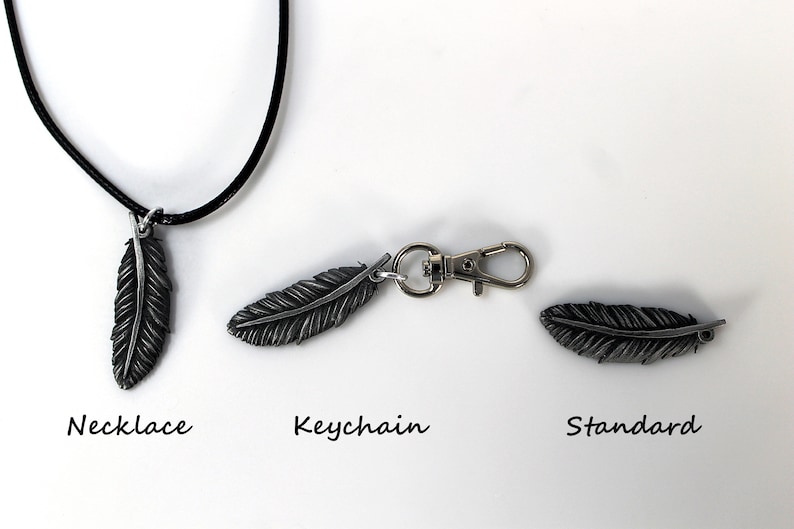Op de afbeelding: Een set van drie metalen veervormige hangers. E&eacute;n is een ketting, &eacute;&eacute;n is een sleutelhanger en de derde is een standaard hanger. De veren zijn donker zilverkleurig en hebben gedetailleerde adering.