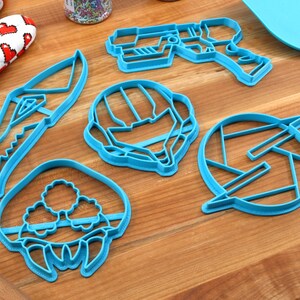 May include: Five blue plastic cookie cutters shaped like a space helmet, a gun, a creature, a knife, and a symbol.