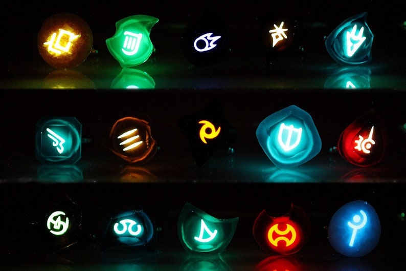 May include: A set of 15 glowing resin rings with different symbols and colors. The rings are arranged in three rows of five. The symbols include a sword, a shield, a fire, a water, a wind, and a earth. The colors include blue, green, red, yellow, and orange.