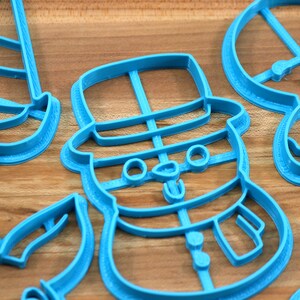 Op de afbeelding: Een set van vier blauwe plastic koekjesvormen in de vorm van een sneeuwpop, een kerstboom, een snoepstok en een rendier. De vormen liggen op een houten oppervlak.