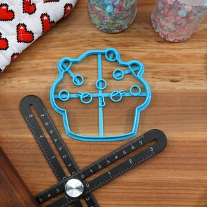 May include: A blue plastic cupcake cookie cutter with a detailed design. The cutter is on a wooden surface with a ruler, a rolling pin, and a blue spatula.