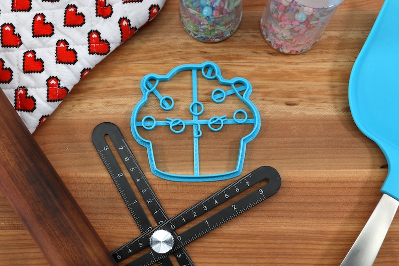 May include: A blue plastic cupcake cookie cutter with a detailed design. The cutter is on a wooden surface with a ruler, a rolling pin, and a blue spatula.