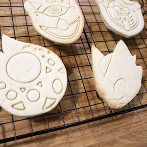 May include: Four sugar cookies on a black wire cooling rack. The cookies are in the shapes of a cat, a flame, a hedgehog, and a sword. The cookies are light brown in color and have white icing.