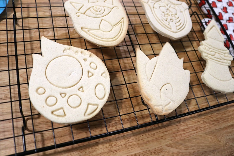 May include: Four sugar cookies on a black wire cooling rack. The cookies are in the shapes of a cat, a flame, a hedgehog, and a sword. The cookies are light brown in color and have white icing.