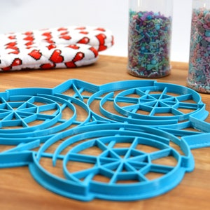May include: A blue plastic cookie cutter shaped like a snowflake with three concentric circles. The cutter is on a wooden surface.