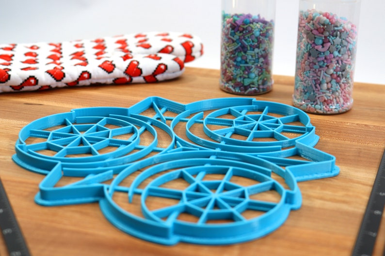 May include: A blue plastic cookie cutter shaped like a snowflake with three concentric circles. The cutter is on a wooden surface.