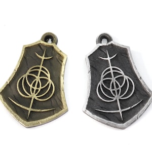 May include: Two metal pendants with a shield shape and a geometric design. The pendant on the left is gold and the pendant on the right is silver.