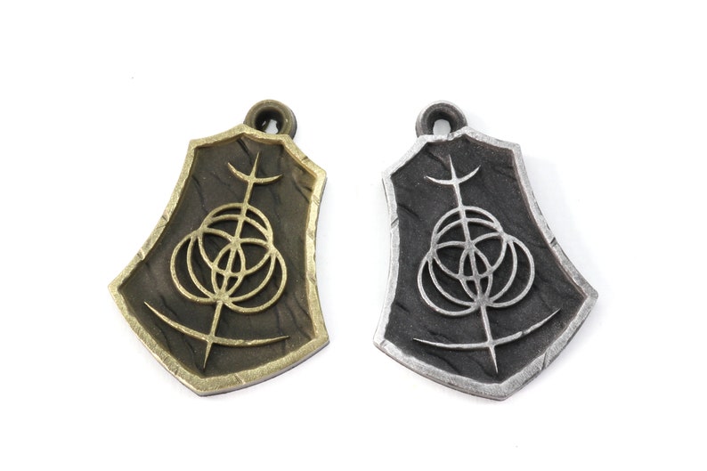 May include: Two metal pendants with a shield shape and a geometric design. The pendant on the left is gold and the pendant on the right is silver.