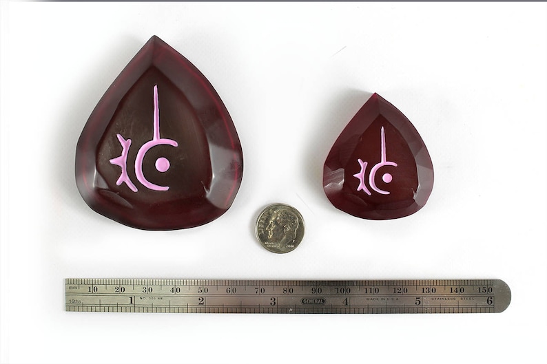 Op de afbeelding: Twee traanvormige, donkerrode glazen bedels met een roze symbool op elk. Het symbool is een gestileerd oog met een halve maan en een verticale lijn.