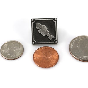 May include: A silver-toned metal pin with a stylized fish design. The pin is square shaped and has a black background. The fish is silver and is outlined in black. The pin is about the size of a US quarter.