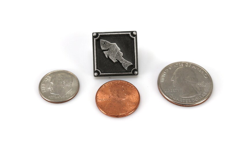 May include: A silver-toned metal pin with a stylized fish design. The pin is square shaped and has a black background. The fish is silver and is outlined in black. The pin is about the size of a US quarter.