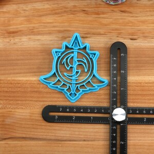 May include: A blue plastic cookie cutter shaped like a stylized symbol with a circular design and a central point. The cutter is approximately 2 inches in diameter.