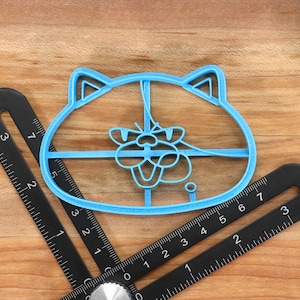 May include: A blue plastic cookie cutter shaped like a cat's face. The cutter is about 5 inches wide and has a detailed design of a cat's face with ears, eyes, nose, and mouth.