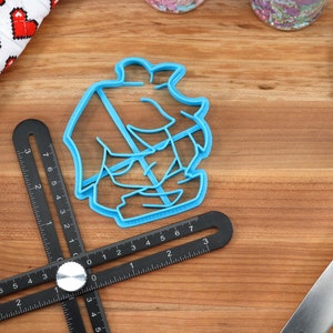 May include: A blue plastic cookie cutter shaped like a ship with a sail. The cutter is on a wooden surface with a black ruler.