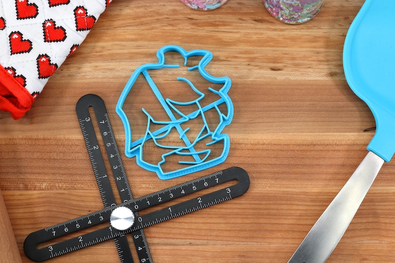 May include: A blue plastic cookie cutter shaped like a ship with a sail. The cutter is on a wooden surface with a black ruler.