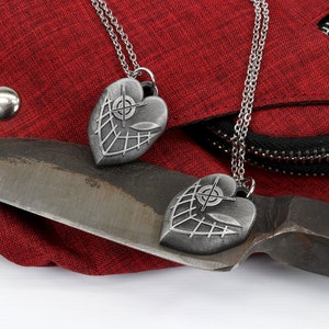 May include: Two silver heart-shaped pendants with a crosshair design and a web-like pattern on a red fabric background. The pendants are hanging from silver chains.