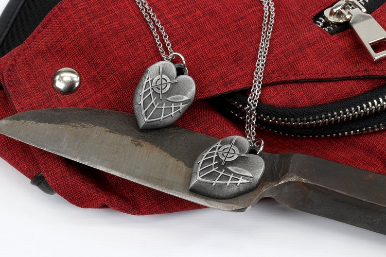 May include: Two silver heart-shaped pendants with a crosshair design and a web-like pattern on a red fabric background. The pendants are hanging from silver chains.