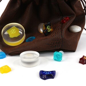 May include: A brown leather dice bag with a drawstring closure, spilling out a variety of colorful dice with different symbols. The dice are made of clear glass with various colors and symbols.