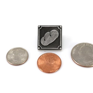 May include: A silver-colored pin with a gray bread design on a black background. The pin is surrounded by a silver border. The pin is next to three US coins: a quarter, a penny, and a dime.