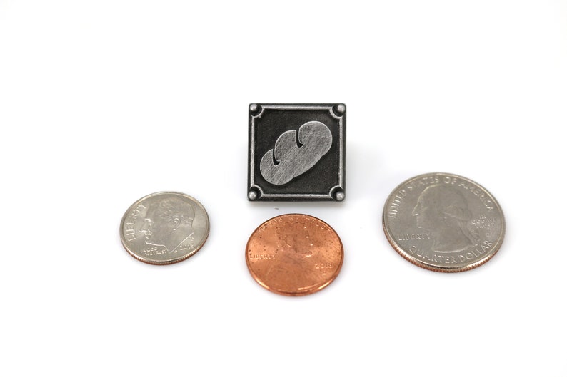 May include: A silver-colored pin with a gray bread design on a black background. The pin is surrounded by a silver border. The pin is next to three US coins: a quarter, a penny, and a dime.
