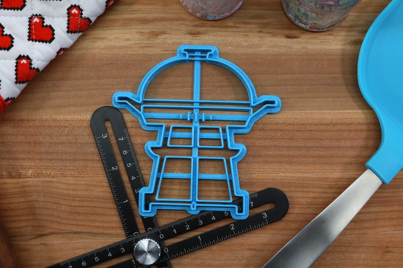 K&ouml;nnte beinhalten: Blauer Plastik-Ausstecher in Form eines Grills. Der Ausstecher liegt auf einer Holzoberfl&auml;che mit einem Lineal und einem blauen Spatel.