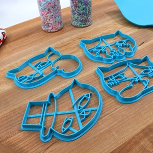 May include: Four blue plastic cookie cutters shaped like cartoon faces with different expressions. The cutters are on a wooden surface.