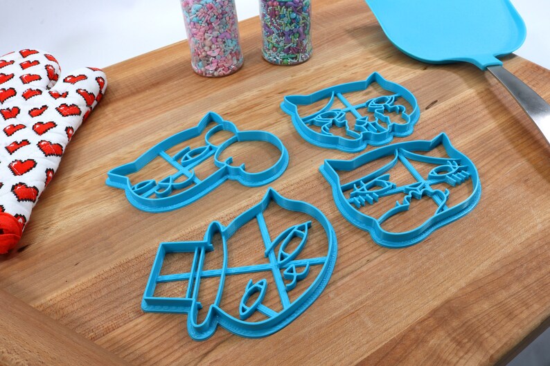 May include: Four blue plastic cookie cutters shaped like cartoon faces with different expressions. The cutters are on a wooden surface.