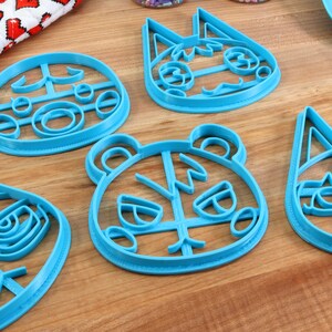 May include: Five blue plastic cookie cutters shaped like animal faces. The cutters are on a wooden surface. The animal faces include a bear, a cat, and a dog.