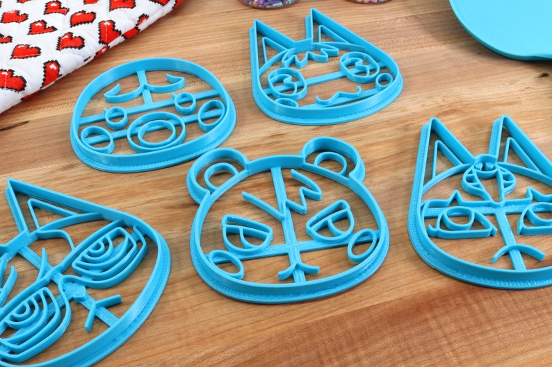 May include: Five blue plastic cookie cutters shaped like animal faces. The cutters are on a wooden surface. The animal faces include a bear, a cat, and a dog.