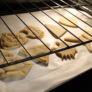 Puede incluir: Una bandeja para hornear con recortes de masa de galletas sin hornear en forma de diamante, estrella, escudo y algunas otras formas. La bandeja est&aacute; en un horno con la puerta ligeramente entreabierta.