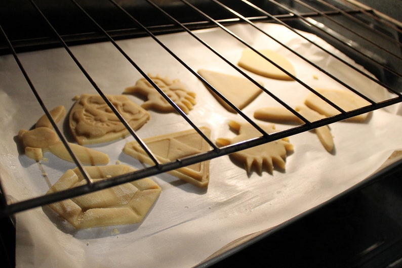 Puede incluir: Una bandeja para hornear con recortes de masa de galletas sin hornear en forma de diamante, estrella, escudo y algunas otras formas. La bandeja est&aacute; en un horno con la puerta ligeramente entreabierta.