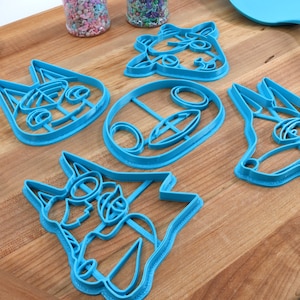 May include: Set of five blue plastic cookie cutters shaped like animal faces. The cutters are on a wooden surface.