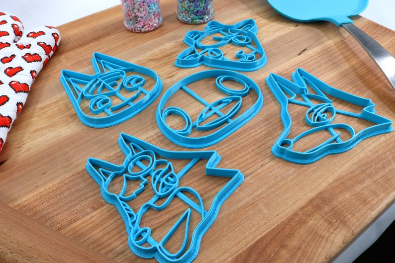 May include: Set of five blue plastic cookie cutters shaped like animal faces. The cutters are on a wooden surface.