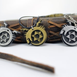 May include: Three metal pendants featuring a stylized dragon design with a star of David inside. The pendants are silver, gold, and bronze in color. The pendants are on a brown leather background.