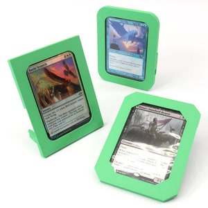 Op de afbeelding: Drie groene kunststof kaarthouders met verschillende Magic: The Gathering kaarten erin. De kaarten zijn "Battle Screech", "Skymarcher" en "Tiana's Standard Bearer".
