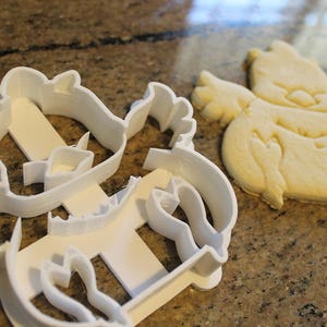 May include: A white plastic cookie cutter in the shape of a cartoon character, next to a baked cookie of the same design. The cookie is light brown and has details pressed into the dough.