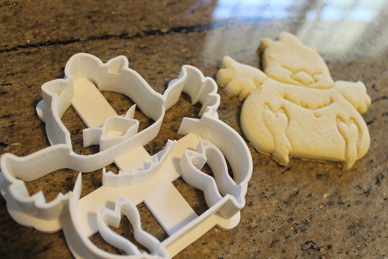May include: A white plastic cookie cutter in the shape of a cartoon character, next to a baked cookie of the same design. The cookie is light brown and has details pressed into the dough.