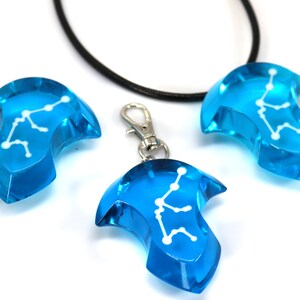 May include: Three blue glass pendants with a white constellation design. The pendants are shaped like a teardrop with a pointed tip. One pendant is on a black cord necklace, and the other two are on silver keychains.