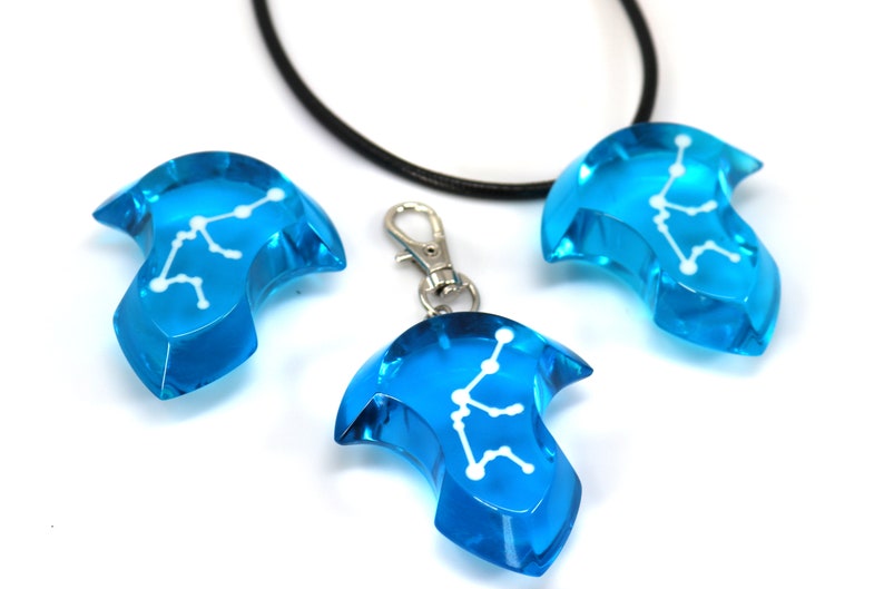 May include: Three blue glass pendants with a white constellation design. The pendants are shaped like a teardrop with a pointed tip. One pendant is on a black cord necklace, and the other two are on silver keychains.