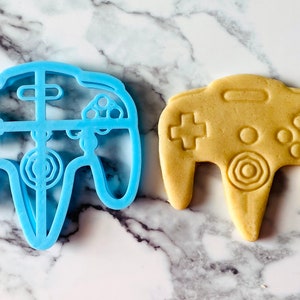 May include: A blue plastic cookie cutter shaped like a Nintendo 64 controller, next to a light brown cookie in the same shape.