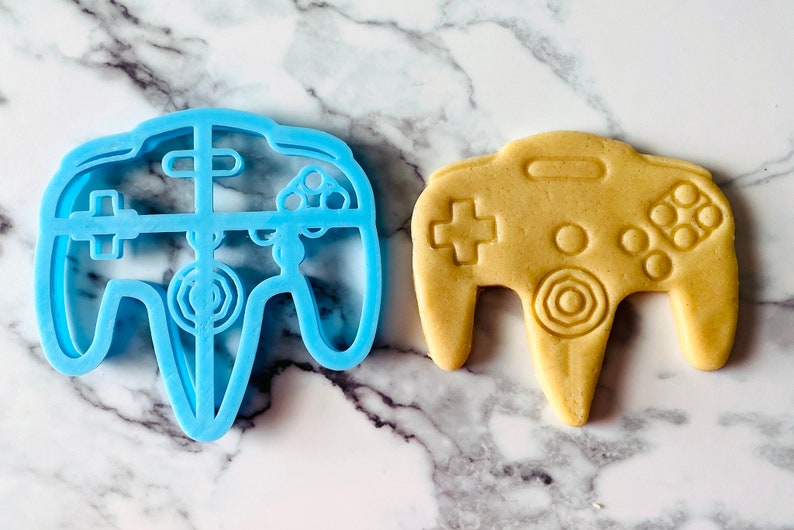 May include: A blue plastic cookie cutter shaped like a Nintendo 64 controller, next to a light brown cookie in the same shape.